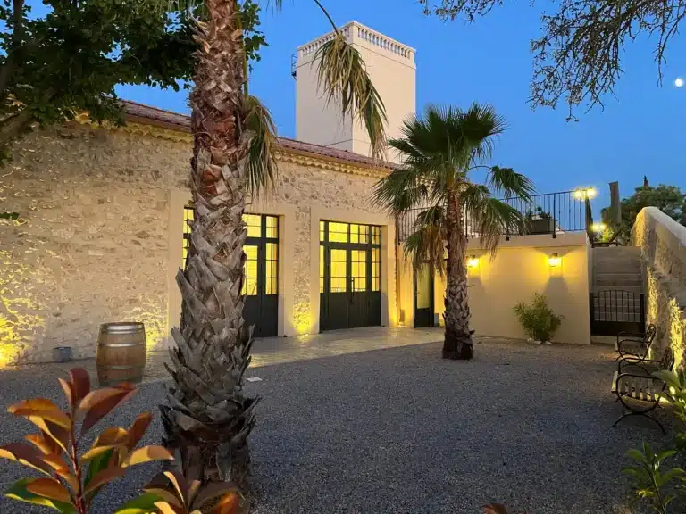 Façade en pierre éclairée avec palmiers, cour extérieure du domaine à Béziers, soirée.